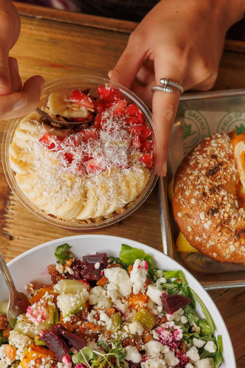 Overhead photo of a woman's hands holding an acai bowl as her spoon is going into it. The acai bowl is near a salad and a breakfast sandwich